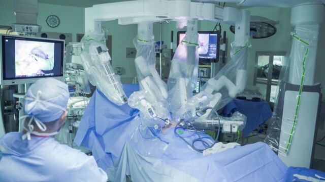 Phoebe Surgeon operates a robotic surgical system in an operating room, with multiple robotic arms covered in plastic and monitors displaying surgical images—her gaming hobby helps her develop the precision needed to help children.